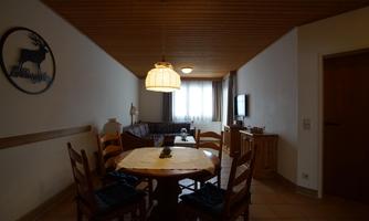 A cozy dining area with a round table and wooden chairs. The walls are bright, and the room has large windows that let in natural light.