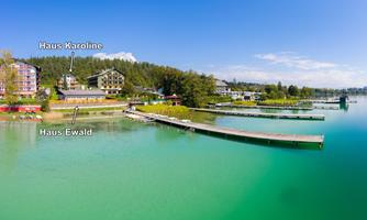 A picturesque lake surrounded by a forested area with two buildings in the foreground. The sky is clear and blue, perfect for a relaxing stay.
