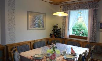 A cozy dining area with a large table set with dishes and decorative napkins. Daylight is shining through the window onto the green garden landscape outside.