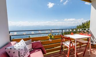 A cozy balcony with a view of the mountains and a bright blue sky. The furniture is simple, with a pink couch and a small table.