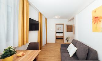 A modern living room with a gray sofa and a wooden table. Large windows with yellow curtains provide plenty of daylight.
