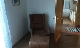 A cozy armchair with a footstool is in a bright corner. In the background, an open kitchen nook is visible.