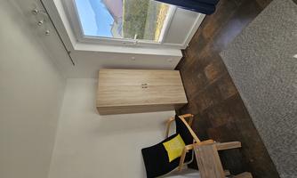 A simple room with a black chair, a small table, and a wardrobe. A window lets in plenty of daylight and offers a view outside.