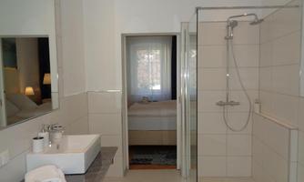 A modern bathroom with a shower and a sink. In the background, a bedroom with a window is visible.