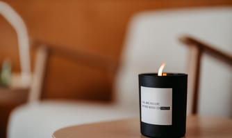 An elegant black scented candle burns on a wooden table. In the background, a modern chair is visible.