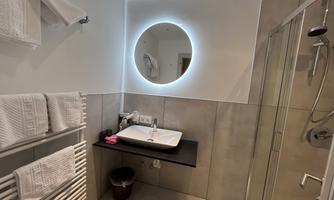 A modern bathroom with a round, illuminated mirror and a sink. Towels hang on a heated towel rail, and a shower is also present.