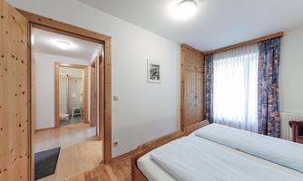 A simple bedroom with two beds and wooden paneling. In the background, a hallway with a bathroom can be seen.