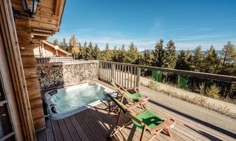 A cozy balcony with a hot tub and two lounge chairs. In the background, tall trees are seen against a clear sky.
