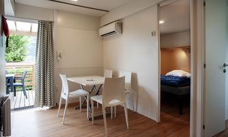 A modern interior with a table and chairs, featuring an air-conditioned bedroom in the background. The room has large windows that let in natural light.