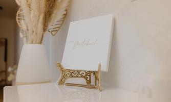 An elegant, minimalist decor with a white book in a gold stand. In the background, decorative plants and a modern vase are visible.