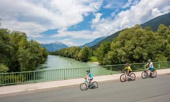 Three cyclists ride along a road with a river in the background. The surroundings are green, and the landscape is characterized by mountains and a clear sky.