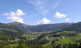 A picturesque landscape with gentle hills and a small village in the valley. The mountains in the background are surrounded by a clear blue sky.