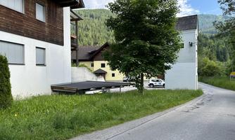 A quiet street with green grass and a small tree. In the background, several buildings can be seen, surrounded by mountains.
