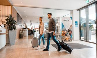 A couple enters the lobby of a hotel with luggage. The modern design and plants give the space an inviting atmosphere.