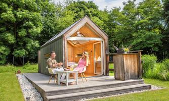 A modern holiday home with a wooden veranda amidst green nature.  
Two people are enjoying their time outside with drinks and nice weather.