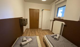A simple bedroom with two single beds and a window. The walls are in neutral colors, and there is a wardrobe as well as towels on the beds.