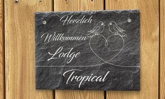 A decorative slate sign with the inscription "Warm Welcome to the Lodge Tropical." It features two birds and a heart motif against a wooden background.