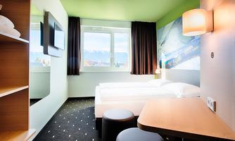 A modern room with two beds, a table, and a television. Large windows provide plenty of daylight and a view of the surroundings.