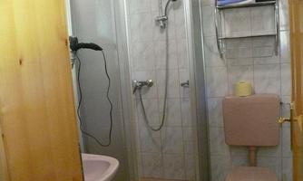 A small bathroom with a shower, a sink, and a toilet. The walls are tiled and there is a shelf with towels.