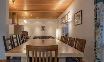 A large dining table with wooden chairs in a cozy room. The ceiling is made of wood, and there is a window with curtains.