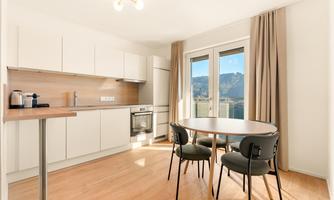 A modern kitchen with a dining table and chairs. Large windows offer a view of the mountains.