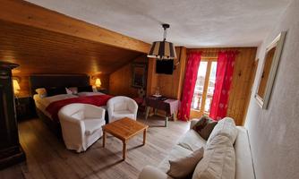 A cozy room with wooden walls and a sloped roof. There are two beds, a small table, and a couch, as well as large windows with red curtains.