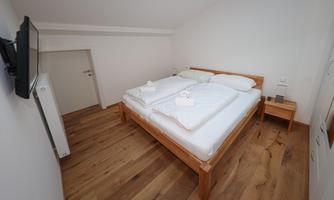 A cozy bedroom with a double bed and wooden floor. A television hangs on the wall, and the walls are painted in a light white.