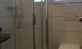 A modern bathroom with a glass shower and a toilet. The walls are tiled in light colors and the window provides natural light.