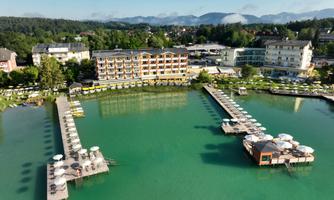 A picturesque lake with clear, green water and several docks. In the background, modern hotels and gentle hills can be seen.