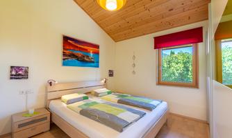 A cozy bedroom with a double bed and colorful bedding. Large windows offer views of the green nature outside.
