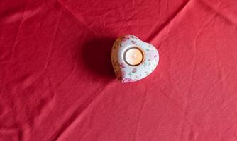 A heart-shaped candle holder with a floral pattern stands on a red table. The candle in the center is lit and emits warm light.