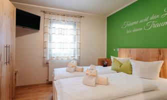 A cozy bedroom with a large window and a television. The walls are painted green, and the bed is neatly made with pillows and blankets.