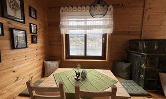 A cozy dining area in a wooden house with a table and chairs. The window offers a view of a picturesque landscape, while decorative pictures adorn the walls.