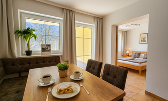 An inviting dining area with a table, two cups of coffee, and a piece of cake. In the background, a bright room with a cozy bed is visible.