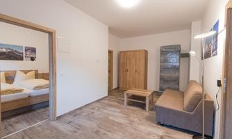 A simple, bright room with wooden flooring and modern furnishings. There is a bed, a wardrobe, and a seating area with a sofa.