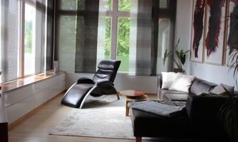 A modern living room with a black reclining chair and a gray sofa. Large windows let in plenty of daylight and offer a view of greenery.