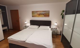 A modern bedroom with a double bed, nightstands, and bedside lamps. A colorful artwork hangs on the wall, and there is a decorative plant in the corner.