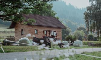 A rustic wooden house stands in a rural setting. In the foreground, there is a tractor and some hay bales.