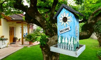 A colorful birdhouse hangs on a tree. In the background, you can see a well-maintained garden and a cozy house.