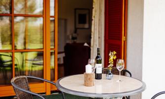 A cozy table on a terrace with a wine, glasses, and a vase of flowers. In the background, large windows and an inviting living space can be seen.