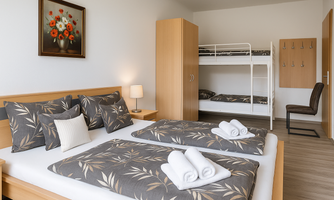 A bright room with two beds, decorated with pillows and towels. In the background, there is a bunk bed and a desk chair.