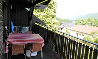 A balcony with a table and chairs. The surroundings are green and some houses can be seen in the background.
