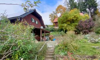 A cozy, red wooden house surrounded by colorful autumn leaves. In the foreground, there is a small garden with many plants and a path.