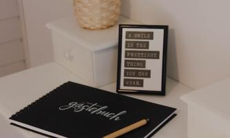 A bright table with a notebook and a pencil. In the background, there is a pot with a plant and a framed quote.