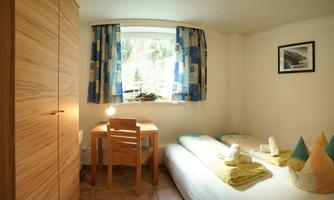 A cozy bedroom with two beds and a small desk. The room has large windows with colorful curtains and minimal furnishings.