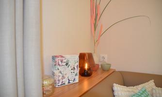 A cozy living area with a wooden shelf, decorated with a flower box, a candle, and a small bowl. In the background, plants and cushions can be seen.