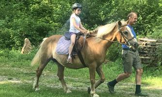 A girl is sitting on a horse while a man leads it. The surroundings are green and friendly.