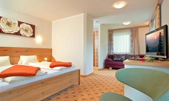 A bright and cozy hotel room with a double bed and a comfortable sofa. There is a television on a table, and there are decorative flowers on the wall.