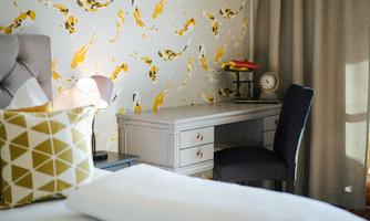 A stylish room with a bed, colorful wallpaper pattern, and a comfortable desk. A cozy atmosphere is created by the soft lighting and decorative cushions.