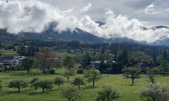 A green landscape with blooming trees and a clear sky, partially covered with clouds. In the background, gentle hills and some residential houses are visible.